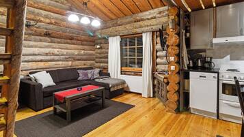 A log cabin interior with a sofa, a coffee table, a kitchen area with appliances, and a wooden ceiling.