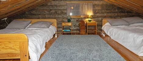 Een gezellige slaapkamer onder de dakpannen met twee houten bedden, een grijs vloerkleed en een klein tafeltje met een lamp.