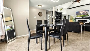 Een moderne eetruimte met een ronde tafel, zwarte stoelen en een kroonluchter. Links staat een televisie op een standaard, en op de achtergrond is een open haard met een bank te zien.