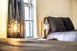 A neatly made bed with a gray comforter and two dark pillows, a bedside table with a lamp, and a window with black curtains.