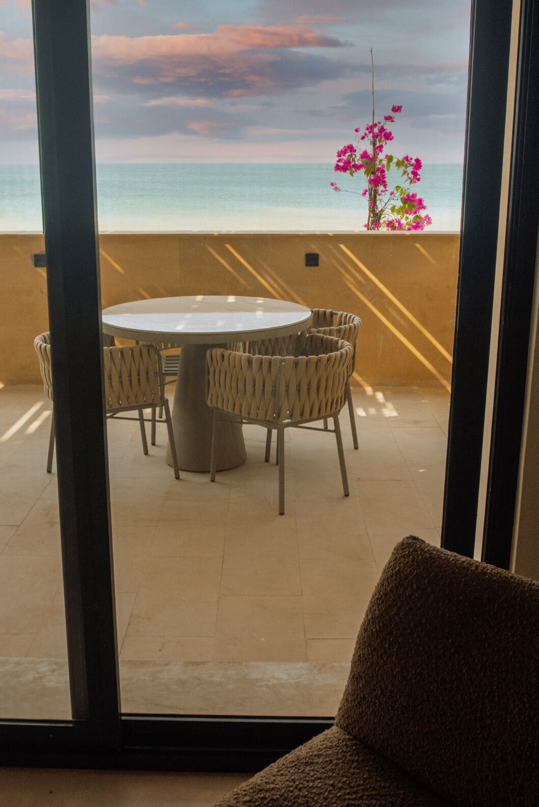 A balcony with a round table and wicker chairs, overlooking a beach with pink flowers.