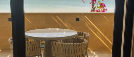 A balcony with a round table and wicker chairs, overlooking a beach with pink flowers.