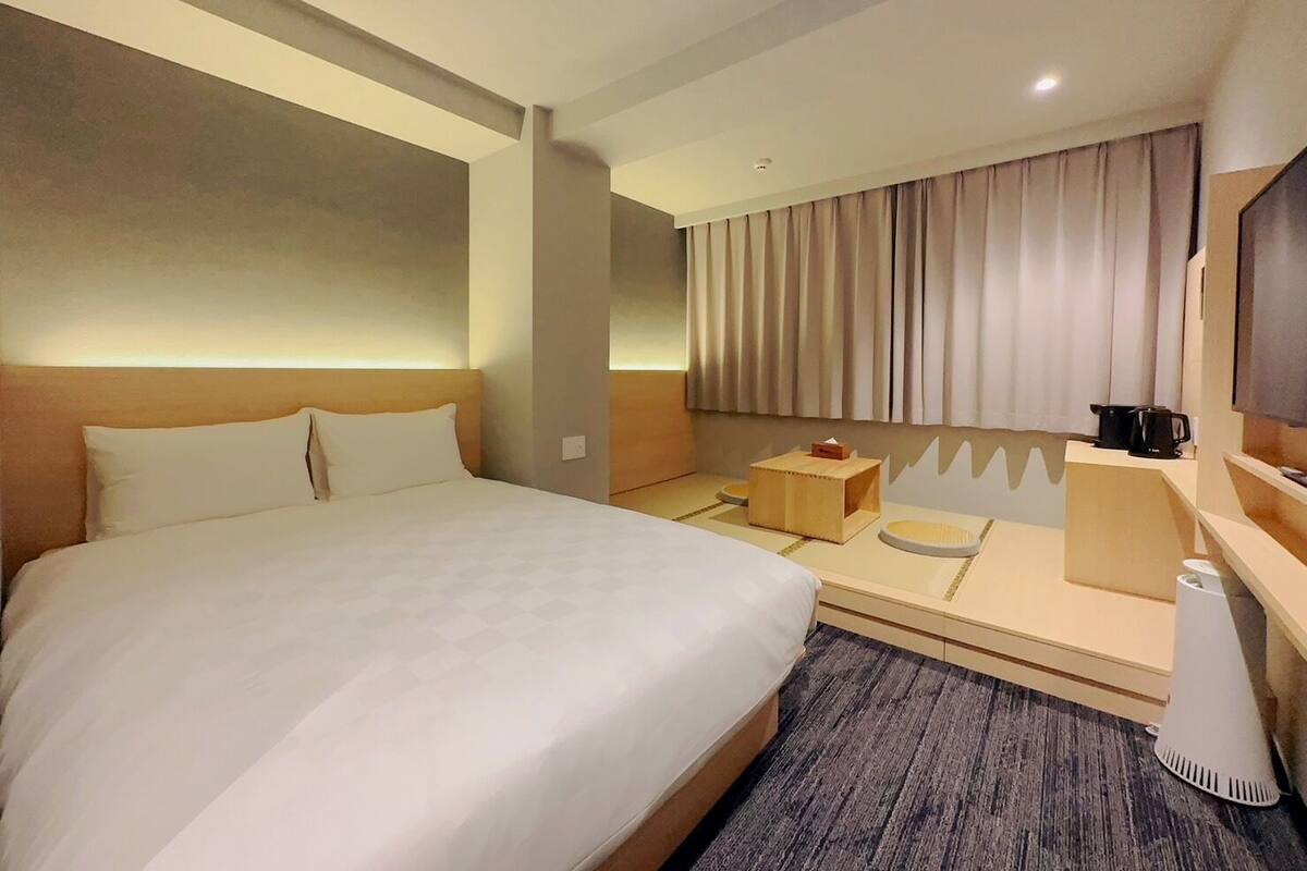 A hotel room with a large bed, a small table with a coffee maker, and a TV on the wall.