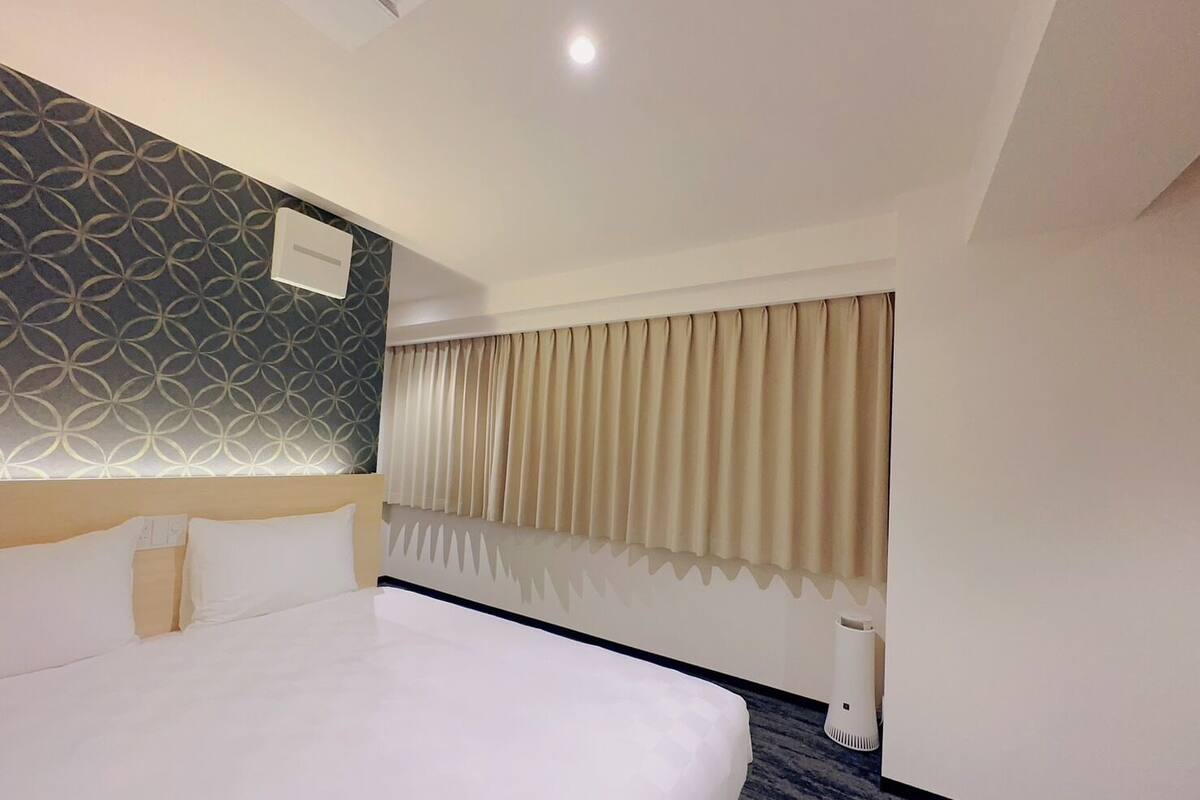 A hotel room with a large bed, a patterned wall, a window with curtains, and a ceiling light.