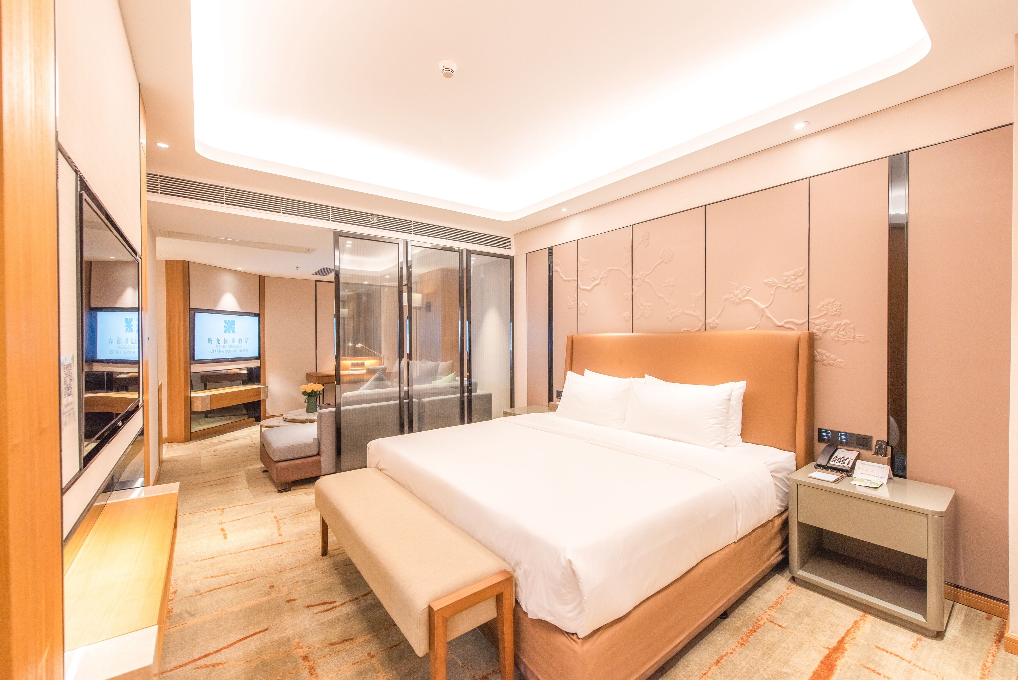 Una habitación de hotel moderna con una cama grande, una mesita de noche con lámpara, un banco, un televisor y una puerta corredera de cristal que da a un baño.