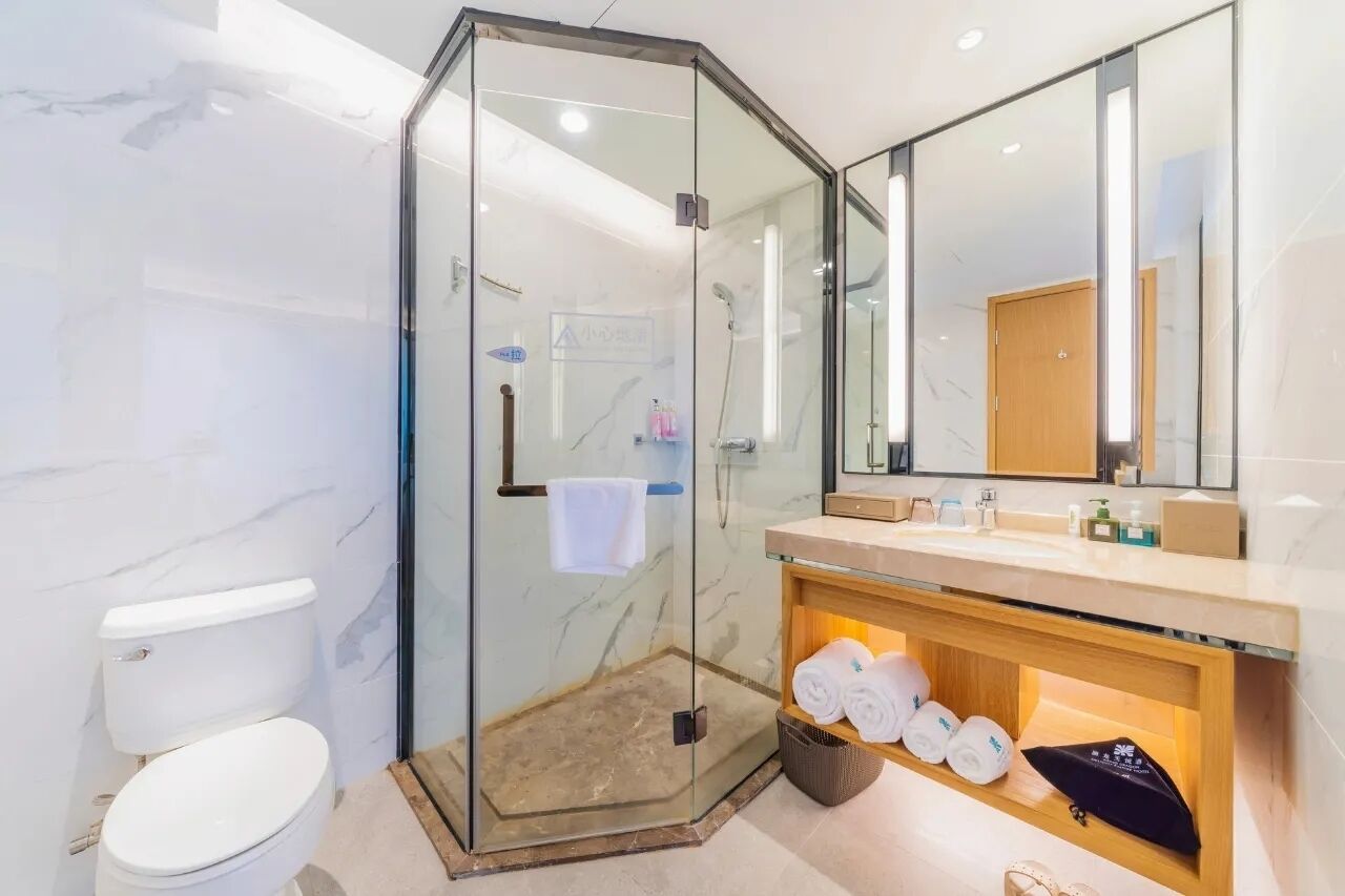 A modern bathroom with a glass-enclosed shower, a marble wall, a white toilet, a vanity with a mirror, and neatly folded towels on a shelf.
