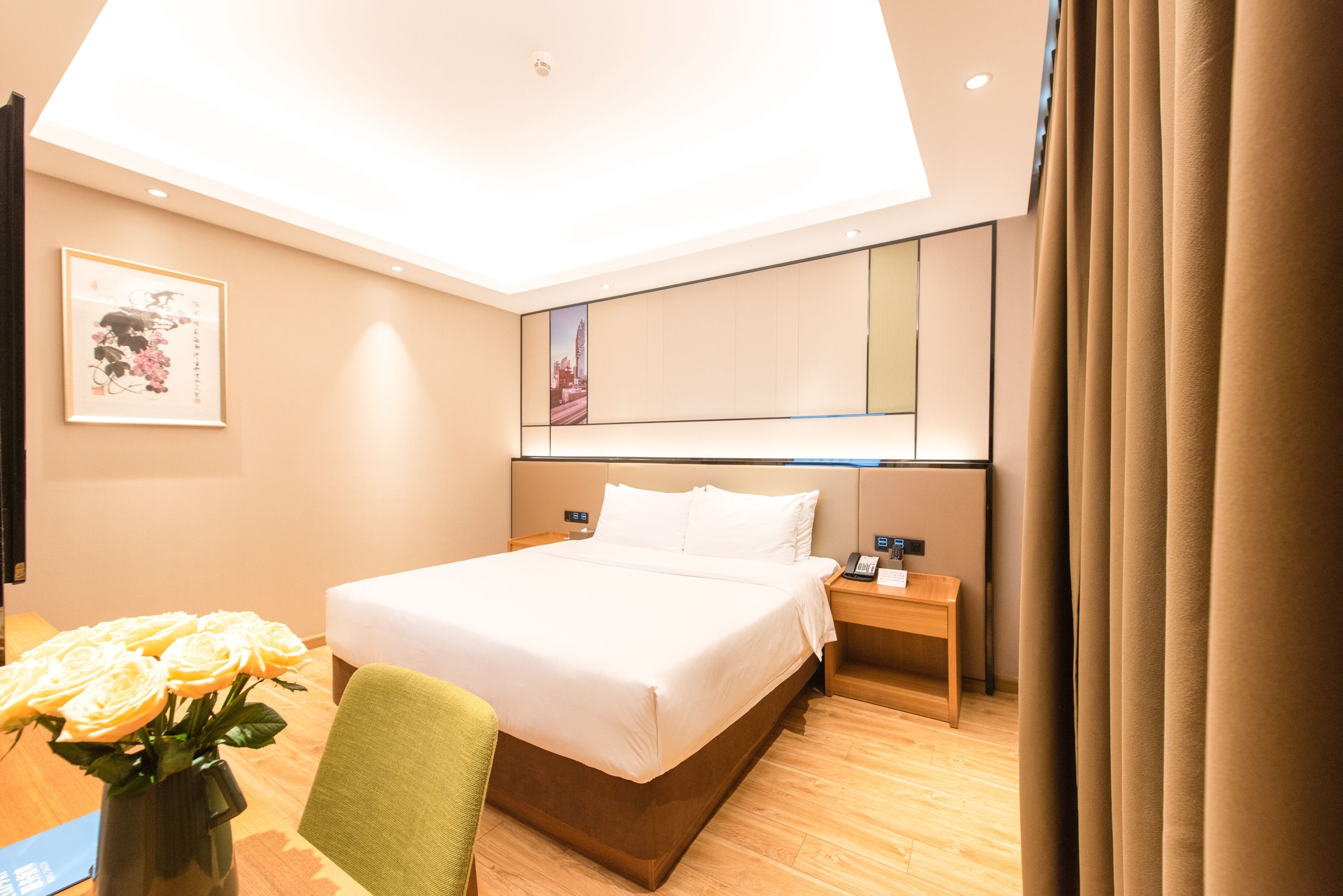 A hotel room with a large bed, bedside tables, a desk with a vase of flowers, and a view of the city through the window.