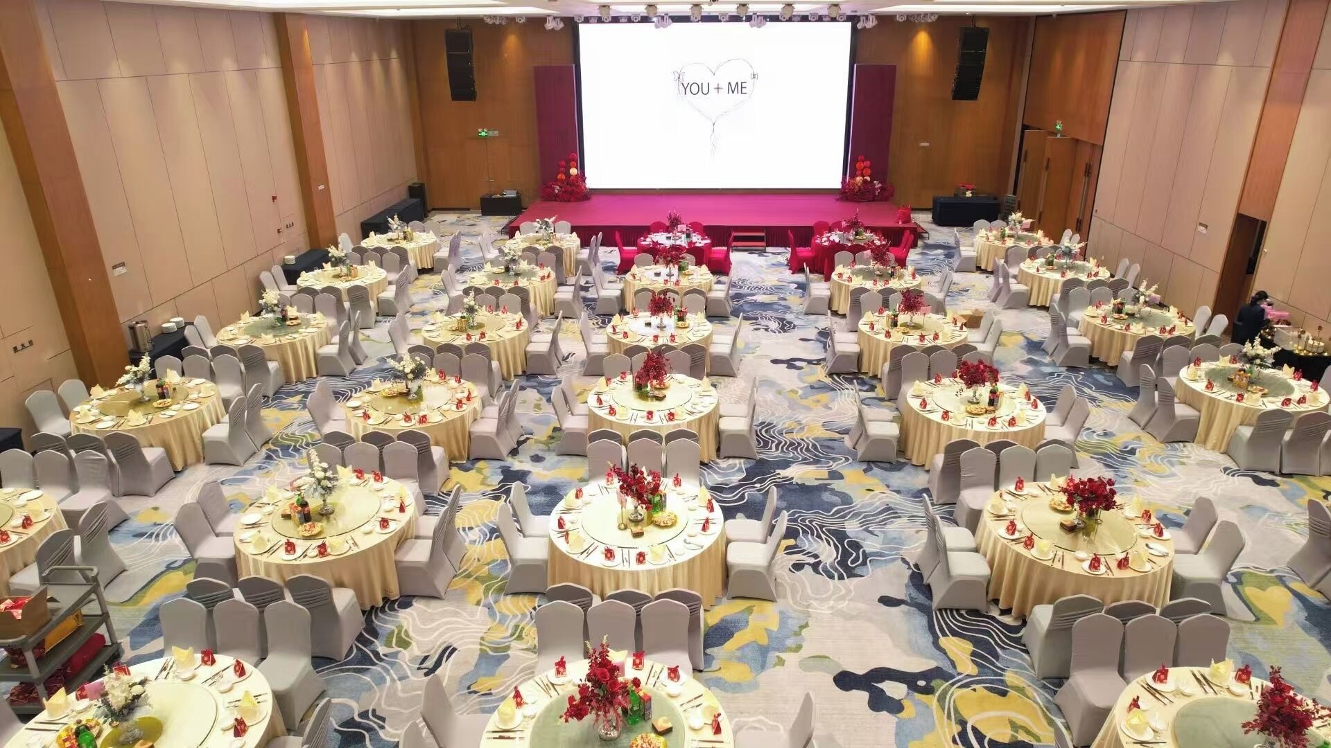A large banquet hall with round tables set for a formal event, a stage with a projection screen, and a carpeted floor.