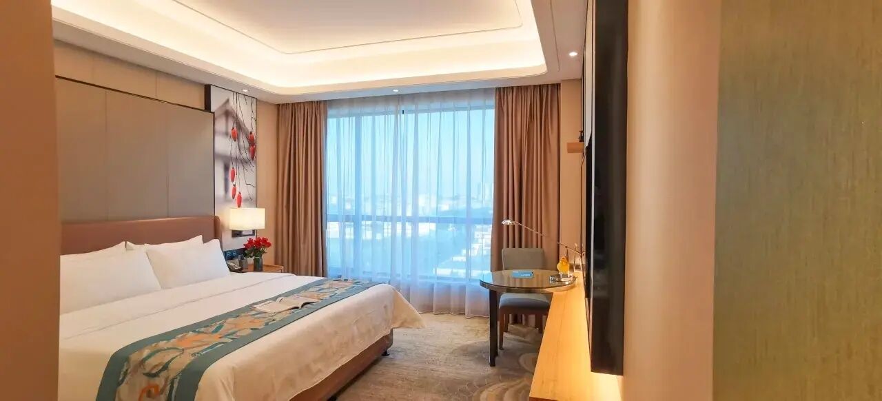 A hotel room with a large bed, a desk, a chair, and a view of the city through the window.