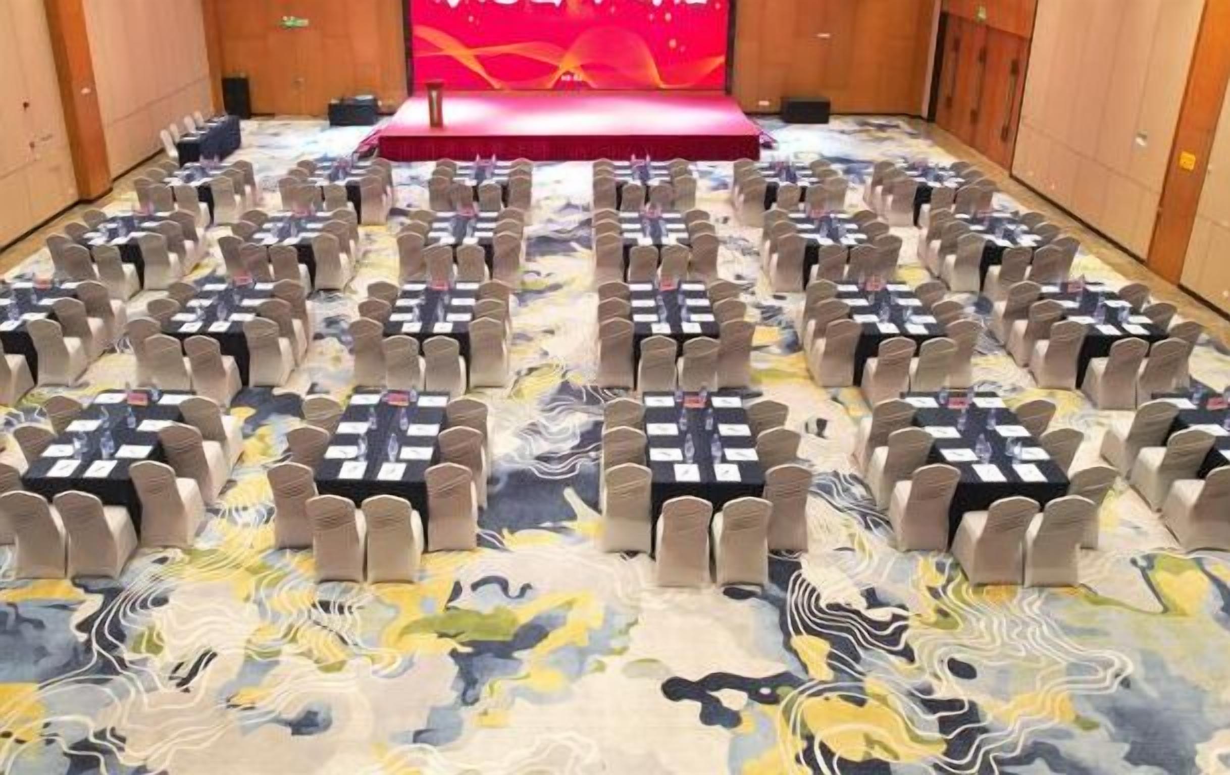 Un salón de conferencias con filas de mesas y sillas, un escenario con un telón rojo y una alfombra estampada.