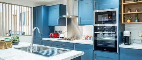 A modern kitchen with blue cabinets, a stainless steel range hood, a built-in oven, and a coffee maker.