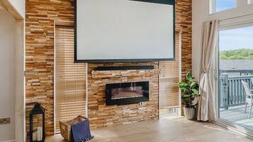 A modern living room with a fireplace, a large TV, wooden wall panels, a balcony with a view, and a wicker basket.