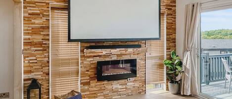 A modern living room with a fireplace, a large TV, wooden wall panels, a balcony with a view, and a wicker basket.