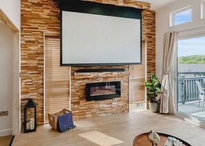 A modern living room with a fireplace, a large TV, wooden wall panels, a balcony with a view, and a wicker basket.