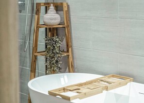 Une salle de bain avec une baignoire blanche, une étagère en bois sur laquelle est posé un vase blanc, et une plante suspendue.