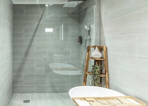 A modern bathroom with grey tiles, a wooden shelf with a white vase, and a bathtub.