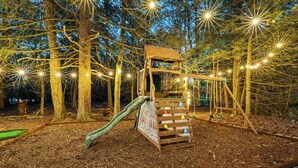 A wooden playground structure with a slide and climbing area, surrounded by trees and illuminated by string lights. - Lakehouse w/ Kayaks, MiniGolf, Sauna, HotTub, Swings and playground for Kids! (Pocono Summit)
