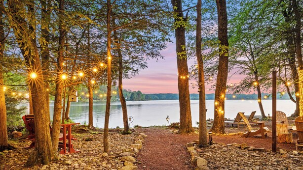 A lakeside path with string lights, wooden chairs, and a bench, leading to a lake. - Lakehouse w/ Kayaks, MiniGolf, Sauna, HotTub, Swings and playground for Kids! (Pocono Summit)