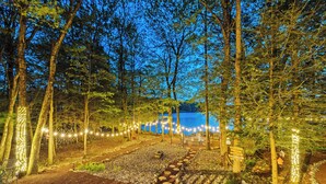 A forested area with a pathway leading to a body of water, decorated with string lights. - Lakehouse w/ Kayaks, MiniGolf, Sauna, HotTub, Swings and playground for Kids! (Pocono Summit)