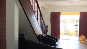 A staircase with wooden railings leading to a bright room with a large window and a fan on the floor.