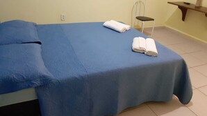 A single-bed room with a blue bedspread, two rolled towels, a chair, and a small table with a lamp.