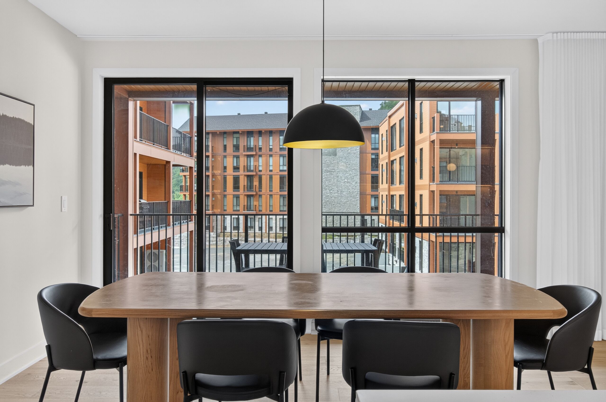 Uma área de refeições moderna com uma mesa grande e cadeiras, uma luminária pendente e vista para um prédio com varandas.