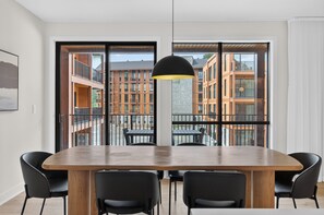 Un espace de salle à manger moderne, doté d’une grande table et de chaises, d’un luminaire suspendu et offrant une vue sur un immeuble à plusieurs étages doté de balcons.