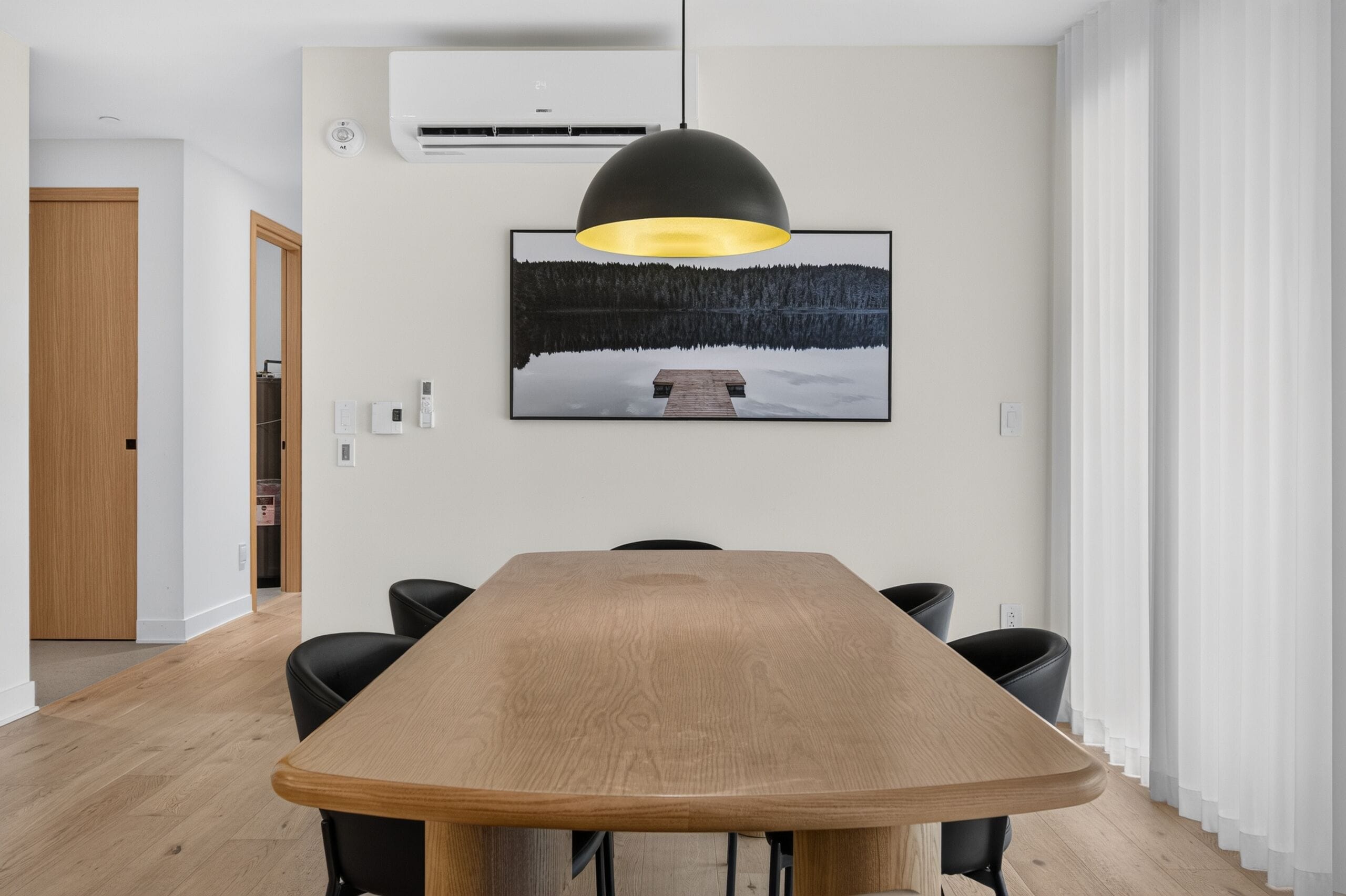 Uma área de refeições com uma mesa de madeira comprida, cadeiras pretas, uma luminária pendente e um quadro na parede.