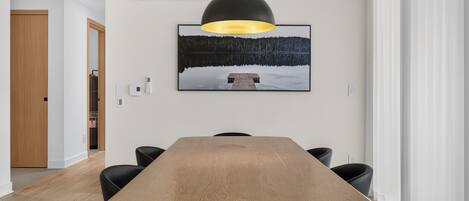 A dining area with a long wooden table, black chairs, a pendant light, and a framed picture on the wall.