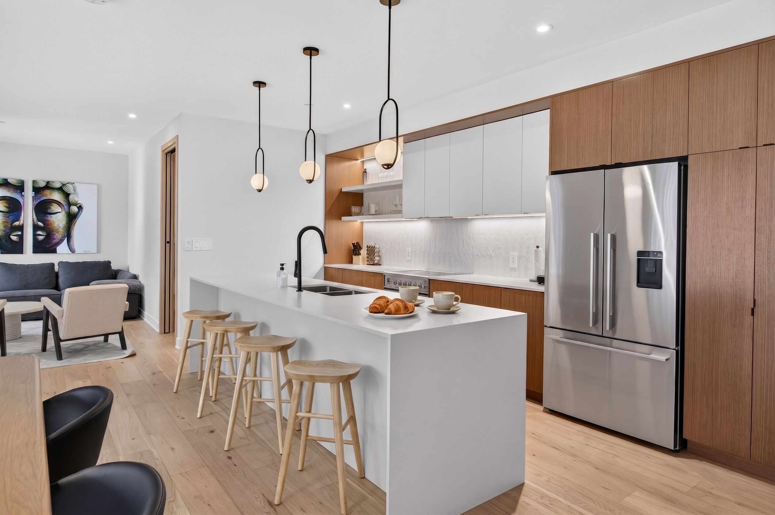 Une cuisine moderne dotée d’un îlot central, de tabourets en bois, d’un réfrigérateur en acier inoxydable et de luminaires suspendus.