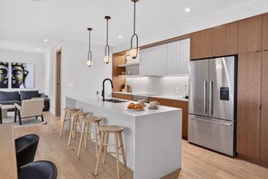 Cozinha moderna com ilha central, banquetas de madeira, geladeira de aço inoxidável e iluminação pendente.