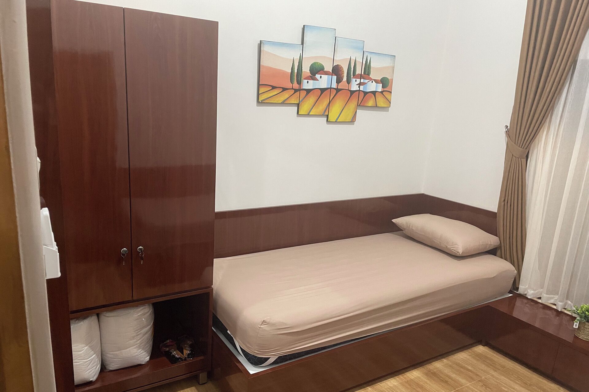 A small, neatly arranged bedroom with a single bed, a wooden wardrobe, and a wall-mounted artwork depicting a landscape.