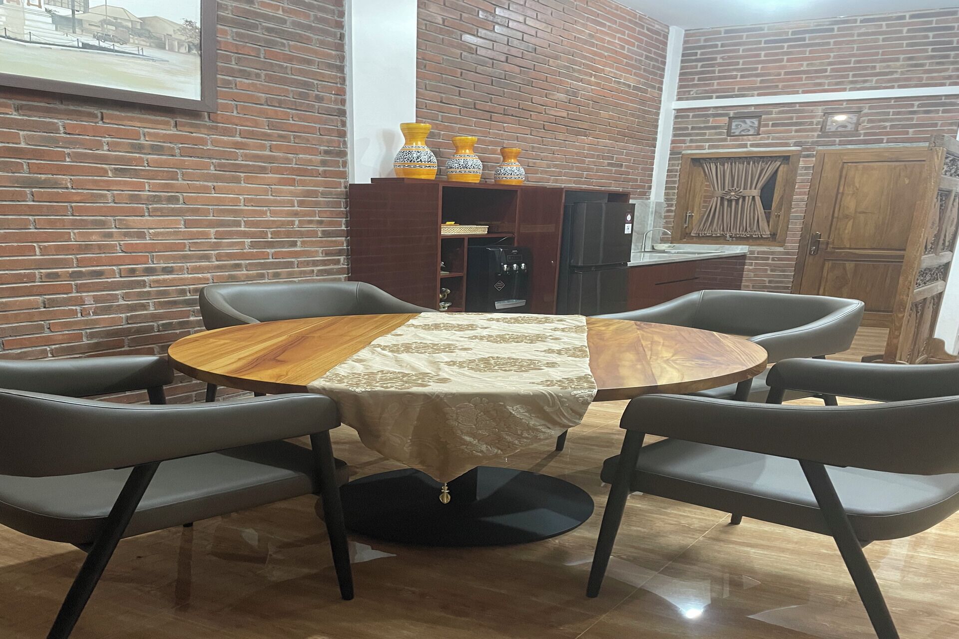 A dining area with a round wooden table, four grey chairs, a coffee machine, and a brick wall.