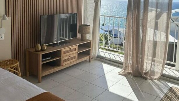 A hotel room with a large TV, wooden panel wall, and a balcony with a view of the sea. - Horizon Caraibes (Le Gosier)