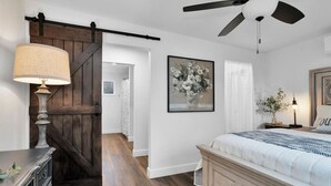 A bedroom with a bed, a nightstand, a lamp, a ceiling fan, and a door leading to another room. - Peaceful Romantic Cottage with Fire Pit and Pond Near Lake Conroe (Montgomery)