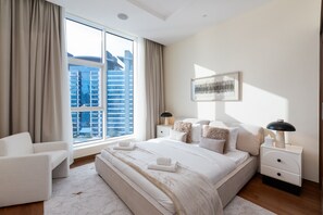 Une chambre à coucher moderne avec une grande fenêtre, un lit recouvert de draps blancs, une table de chevet avec une lampe, et une vue sur des immeubles de grande hauteur.