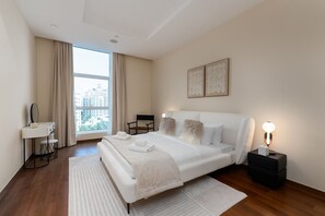 A modern hotel room with a large bed, a bedside table with a lamp, a small desk with a mirror, and a view of the city through the window.