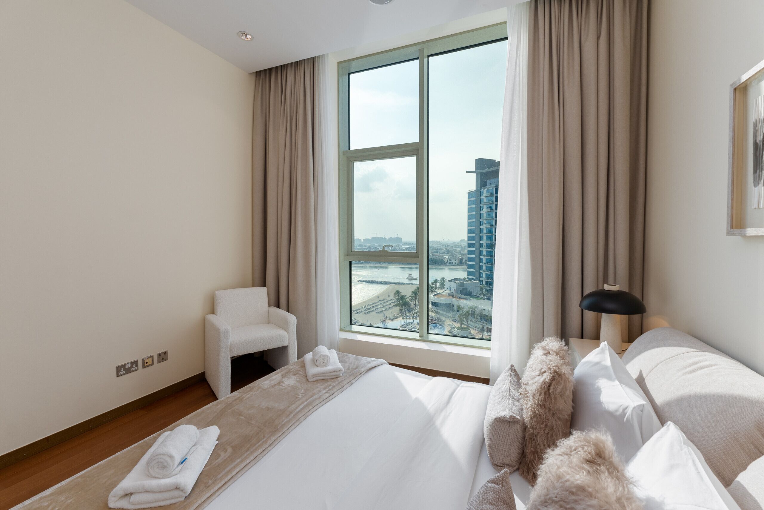 A hotel room with a large window, a bed, a chair, and a lamp.