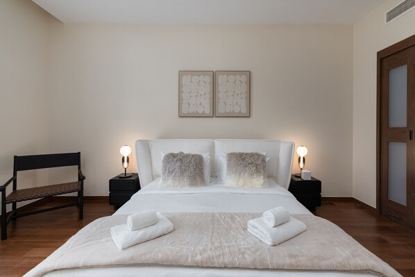 A neatly arranged bedroom with a large bed, two bedside tables with lamps, a wooden bench, and two wall-mounted artworks.
