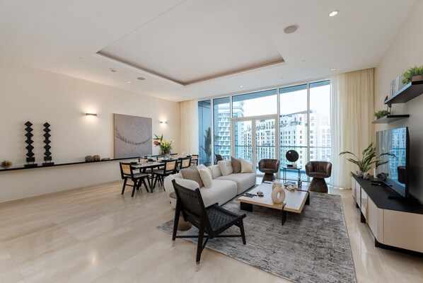 A modern living room with a sofa, coffee table, and chairs, a dining area with a table and chairs, and large windows with a city view.
