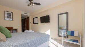 Un dormitorio con cama, ventilador de techo, televisor, espejo y una pequeña mesa con silla.
