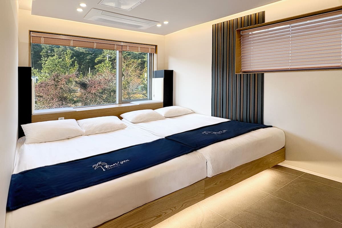 A modern hotel room with a large bed, a window with blinds, and a view of trees.