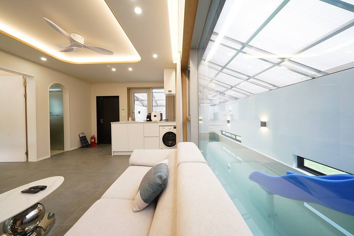 A modern interior with a glass-enclosed swimming pool, a sofa with a pillow, and a kitchen area with a washing machine.