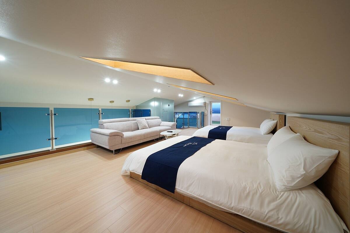 A modern bedroom with a large bed, a sofa, and a view of the sea through a glass door.