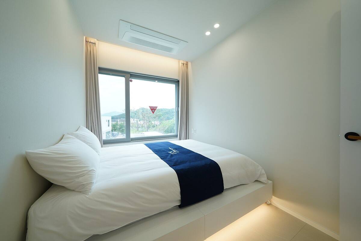 A bedroom with a bed, a window with curtains, a ceiling vent, and a view of a cityscape.