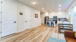 A modern kitchen with white cabinets, a stainless steel refrigerator, and a microwave. There is a wooden floor and a dining area with a bench and chairs.