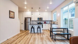 A modern kitchen with white cabinets, a stainless steel refrigerator, a microwave, and a gas stove. There are two black bar stools and a small dining table with benches.