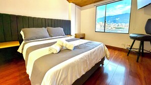 Ein Schlafzimmer mit Bett, Sessel, Nachttisch, Fenster und Blick auf die Stadt sowie einer Holzbalkendecke.