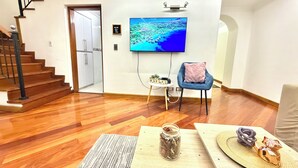 Ein geräumiges Zimmer mit Holzboden, einem blauen Sessel, einem kleinen Tisch mit einem Glas und einem Fernseher an der Wand.