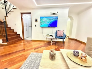 Ein geräumiges Zimmer mit Holzboden, einem blauen Sessel, einem kleinen Tisch mit einem Glas und einem Fernseher an der Wand.